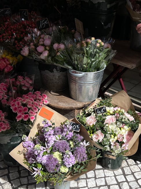 An assortment of fresh flower bouquets displayed outdoors at a florist shop. The arrangement includes a bucket of pink ranunculus flowers with delicate, tightly packed petals and some green stems visi
